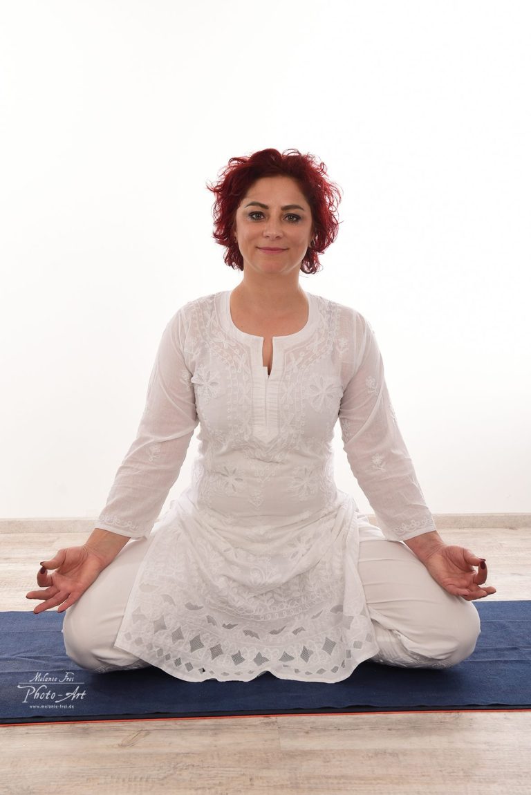Frau in Weiß sitzt im Schneidersitz und meditiert auf einer Yogamatte.