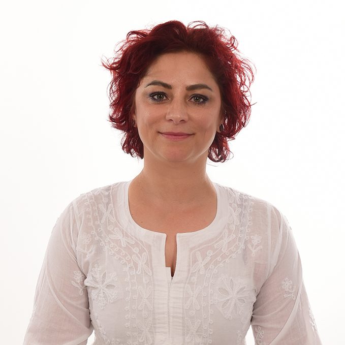 Frau mit roten, lockigen Haaren in einem weißen Oberteil, freundlich lächelnd.